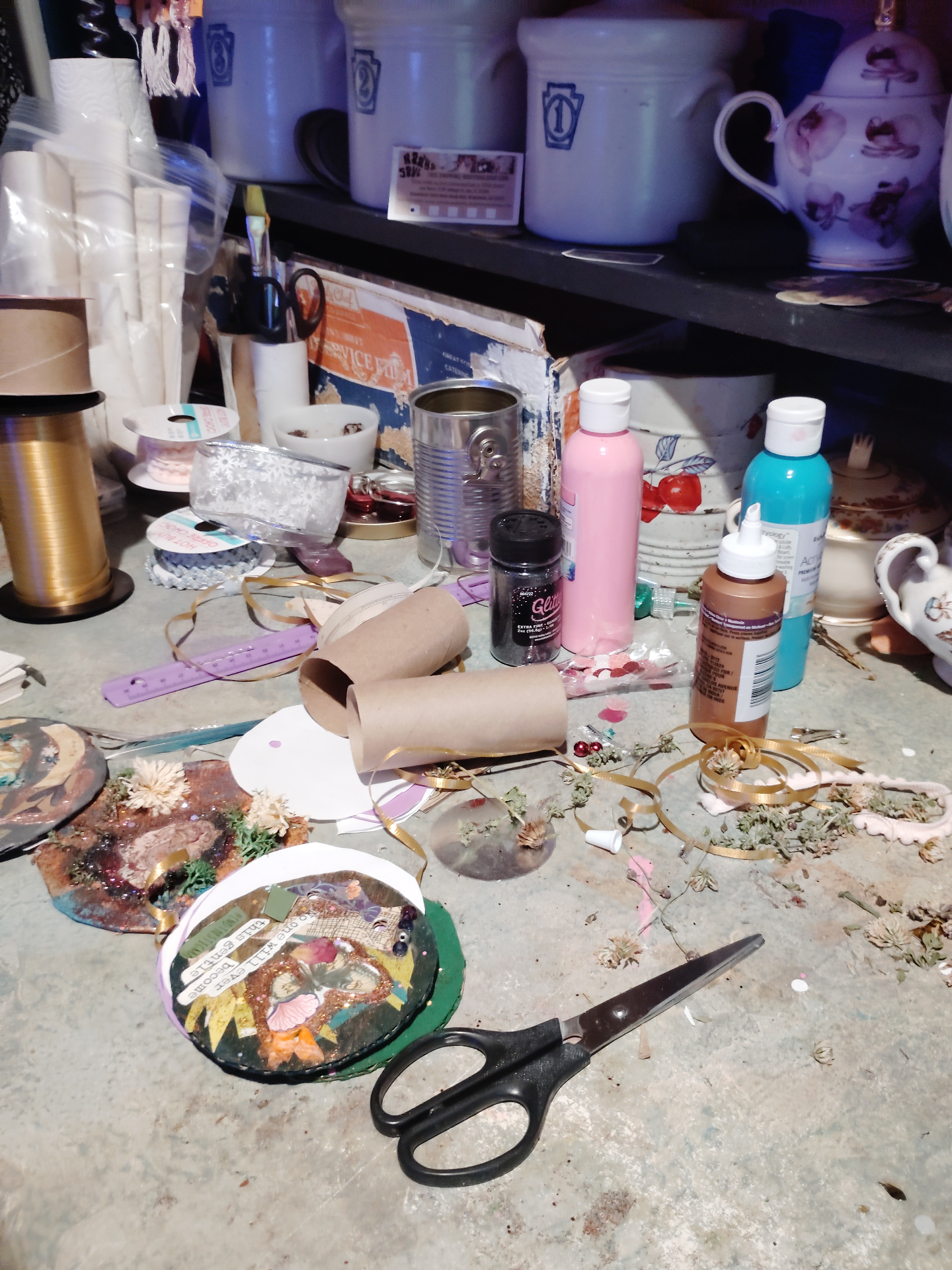 A close view of the worktable with scissors, paint, glitter, ribbon, dried flowers, and handmade mixed-media pieces